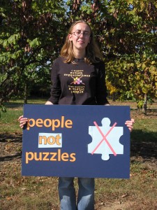 [Image description]A white woman (aspierhetor) with blonde hair, holding a blue sign that reads: "People not puzzles!" (from aspierhetor) 